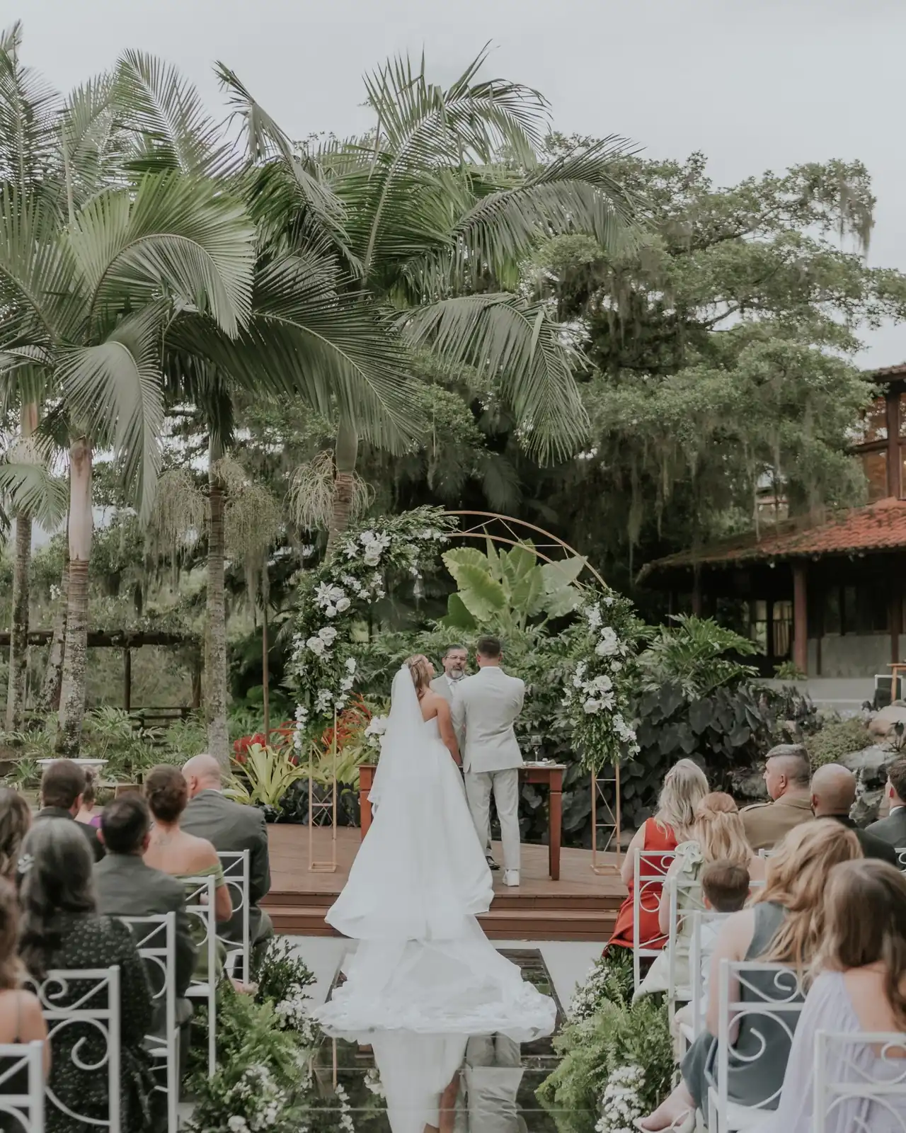 Casal diante de um arco de flores, entre palmeiras e convidados, com o vestido refletido na pista de vidro.