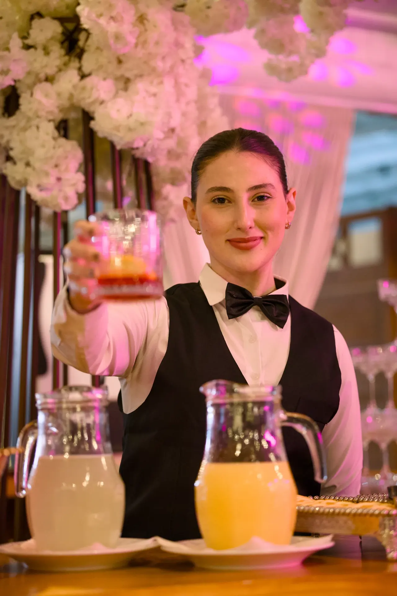 Bartender oferece um drink atrás do bar, entre jarras, flores brancas e luzes da festa.