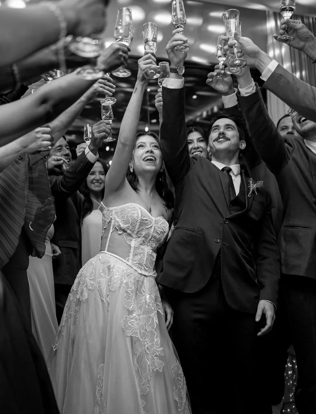 Casal e convidados levantam as taças em um brinde, em uma fotografia em preto e branco.