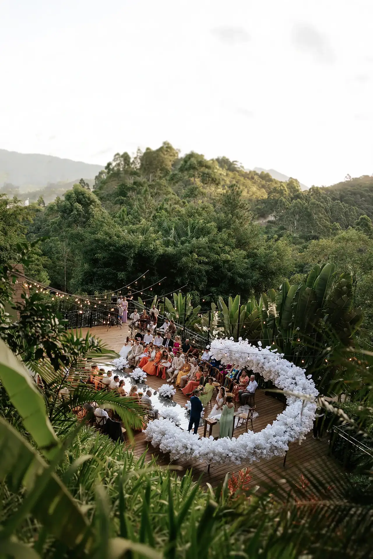 Cerimônia de casamento ao ar livre, com convidados e uma composição de flores brancas, cercada por mata e montanhas sob um céu claro.