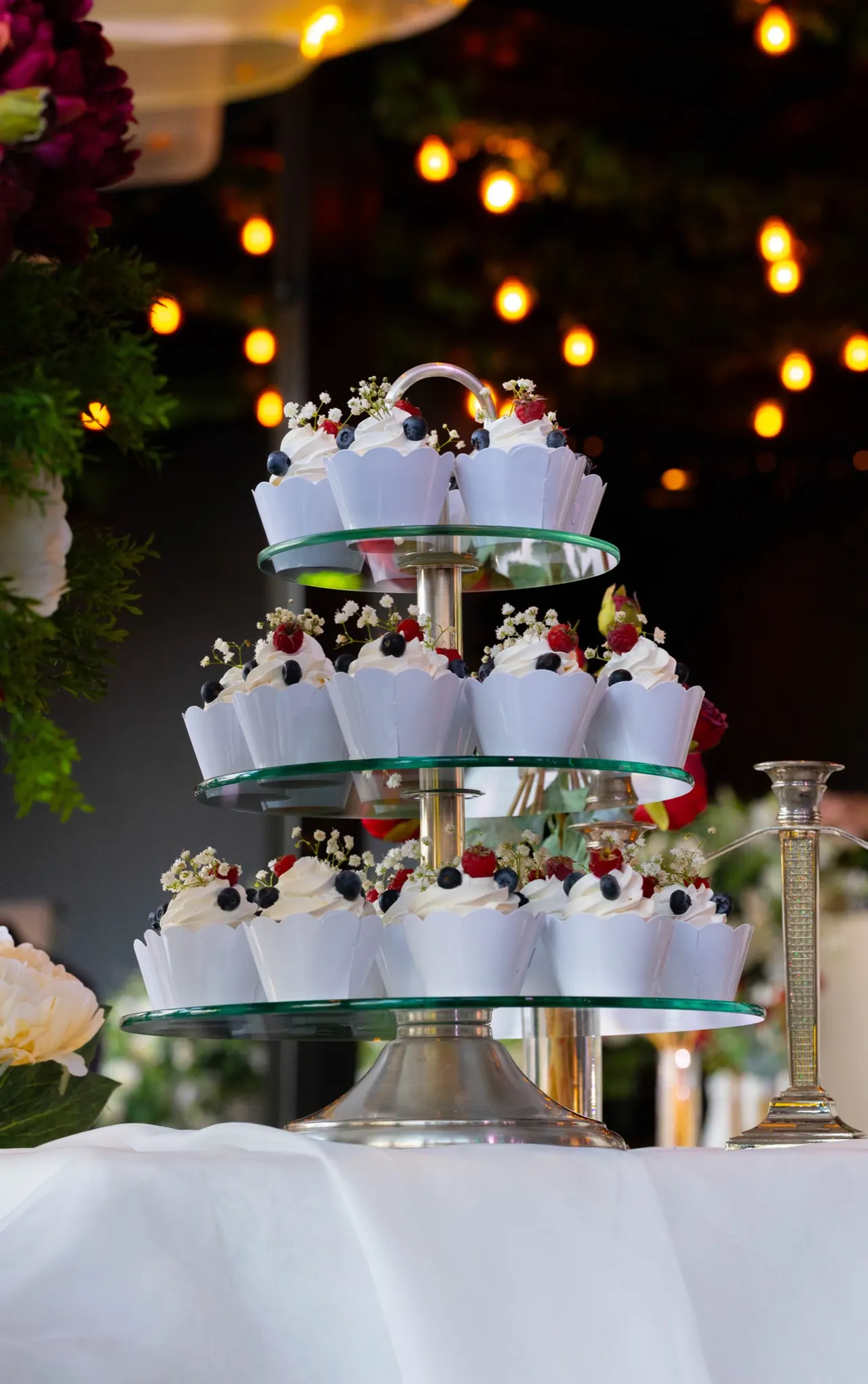 Doces com frutas e flores em uma torre de vidro, com luzes quentes ao fundo.